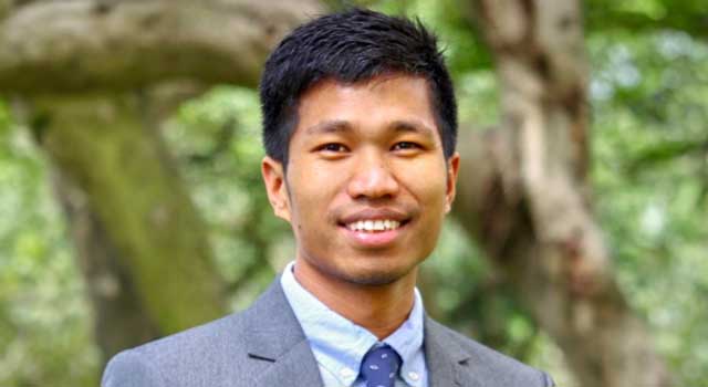 Headshot of a student in a blue tie and grey suit with a plant and nature background